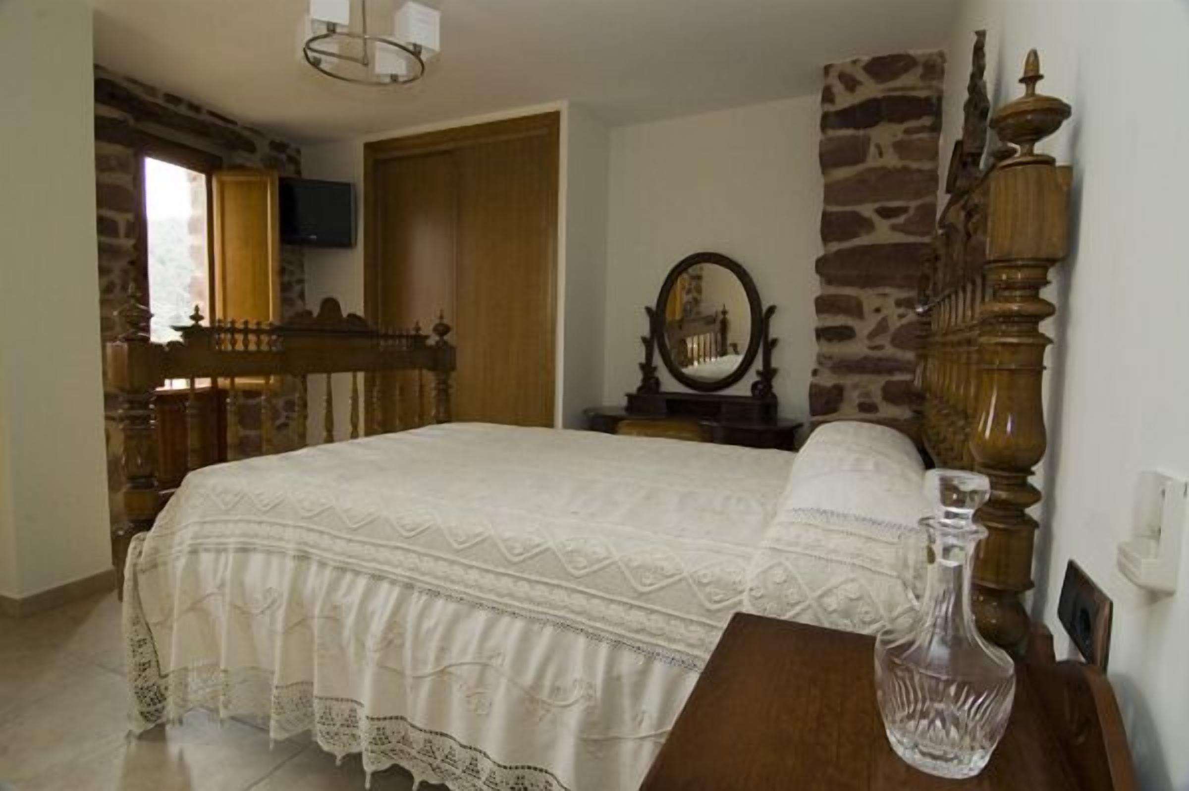 Quarto com cama, mesa de cabeceira de madeira, parede de pedra, espelho e televisão na parede.