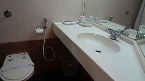 Un baño con lavamanos blanco, espejo e inodoro. Hay un cubo de basura y un dispensador de toallas.