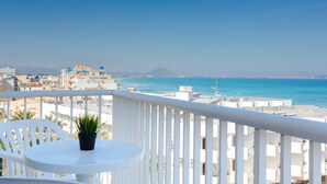 Uma varanda com mesa e cadeira brancas, com vista para um horizonte costeiro com edifícios e o mar.