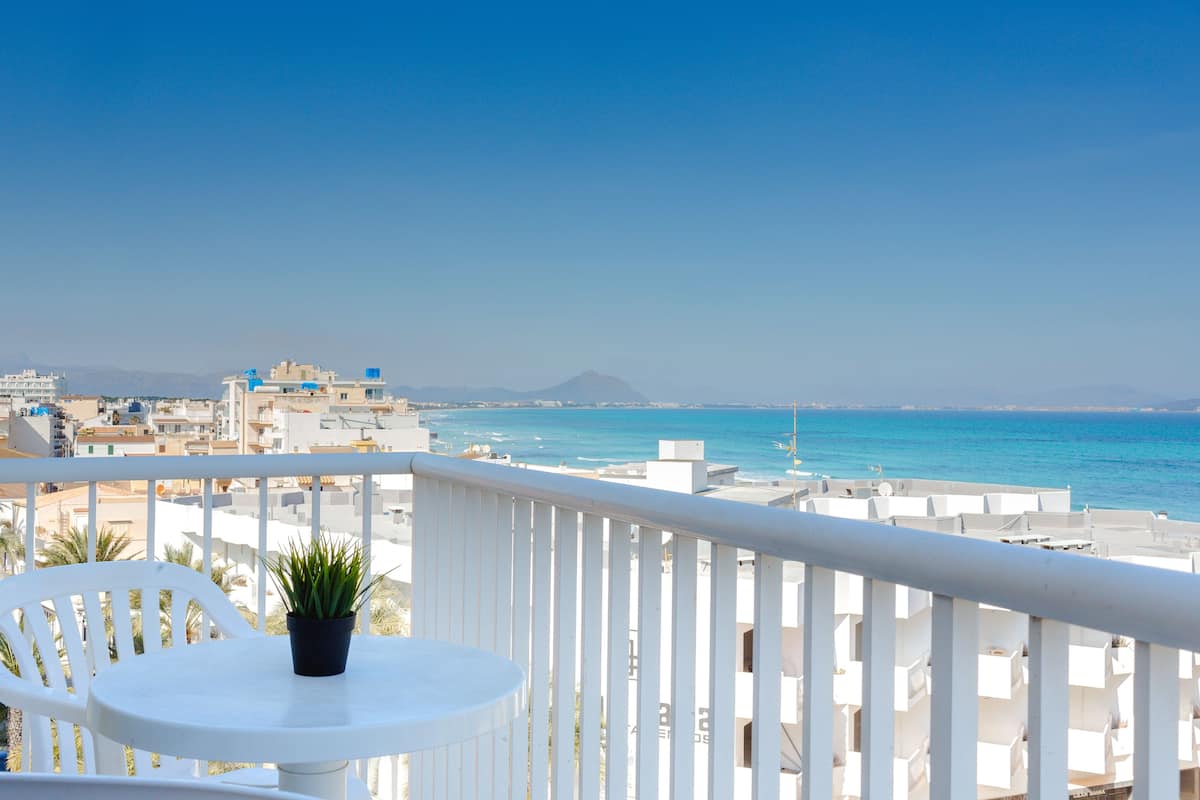 A balcony with a white table and chair, overlooking a coastal cityscape with buildings and the sea.