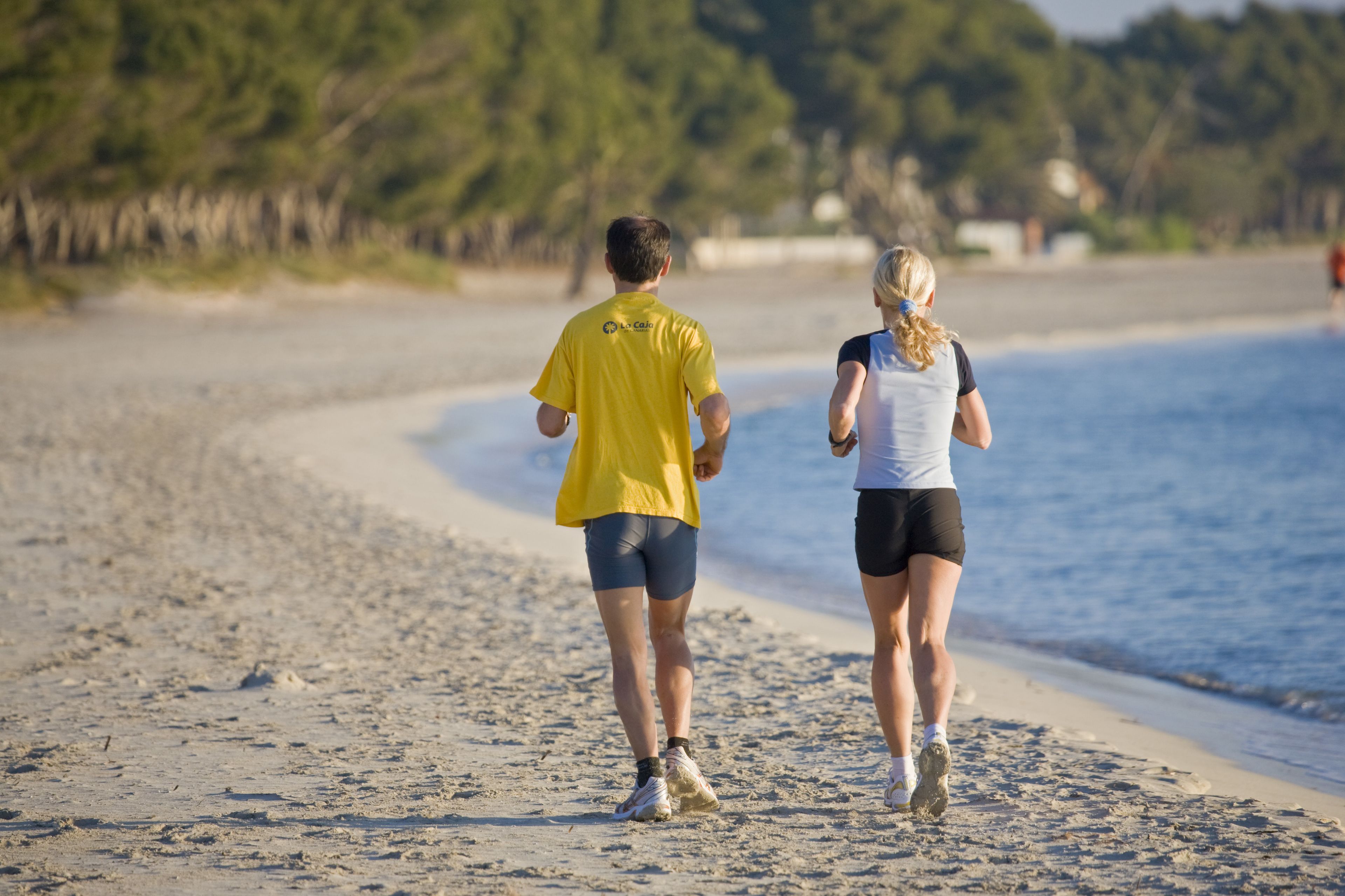 Dos personas caminando sobre una playa arenosa, con una masa de agua y árboles al fondo.