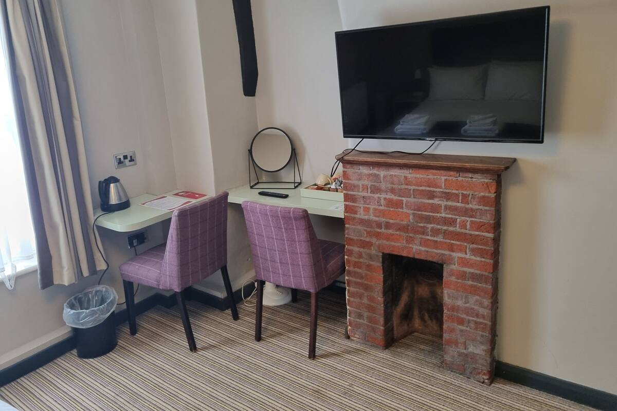 Une chambre d’hôtel avec un téléviseur fixé au mur, une petite cheminée en briques, deux fauteuils, un bureau avec une bouilloire et une poubelle.