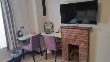 Habitación de hotel con una televisión montada en la pared, una pequeña chimenea de ladrillo, dos sillas, un escritorio con hervidor y un cesto de basura.