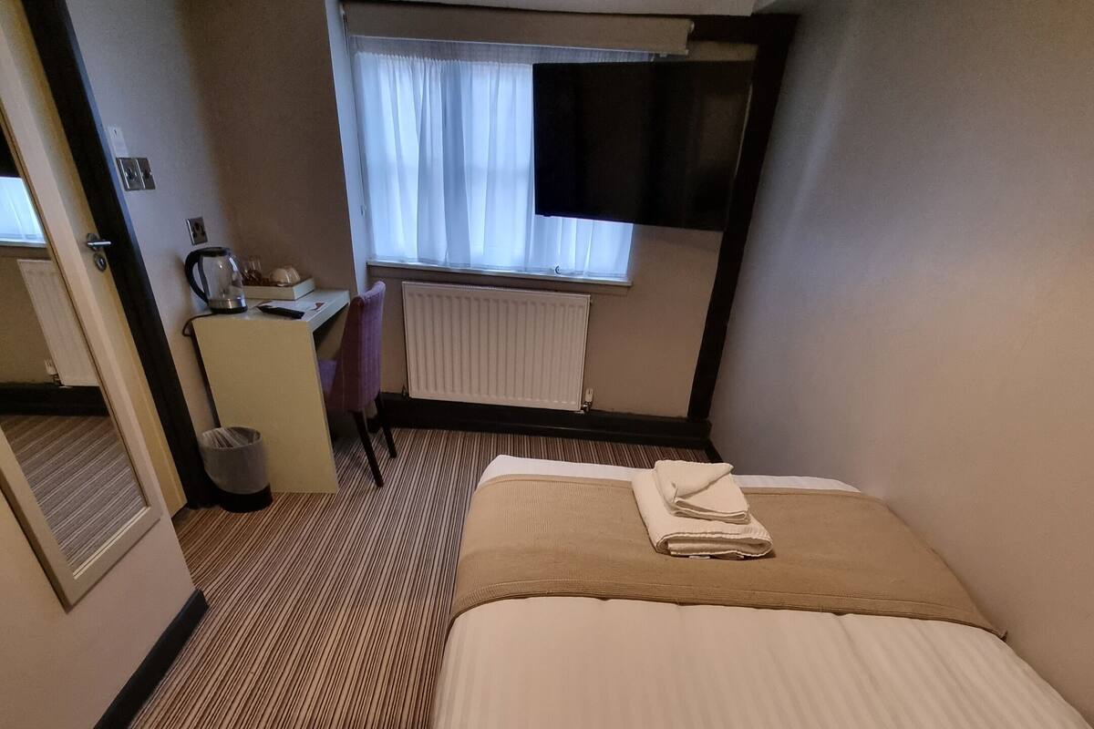 Une chambre d’hôtel avec un lit, un bureau, une chaise, un radiateur et une fenêtre avec des rideaux.