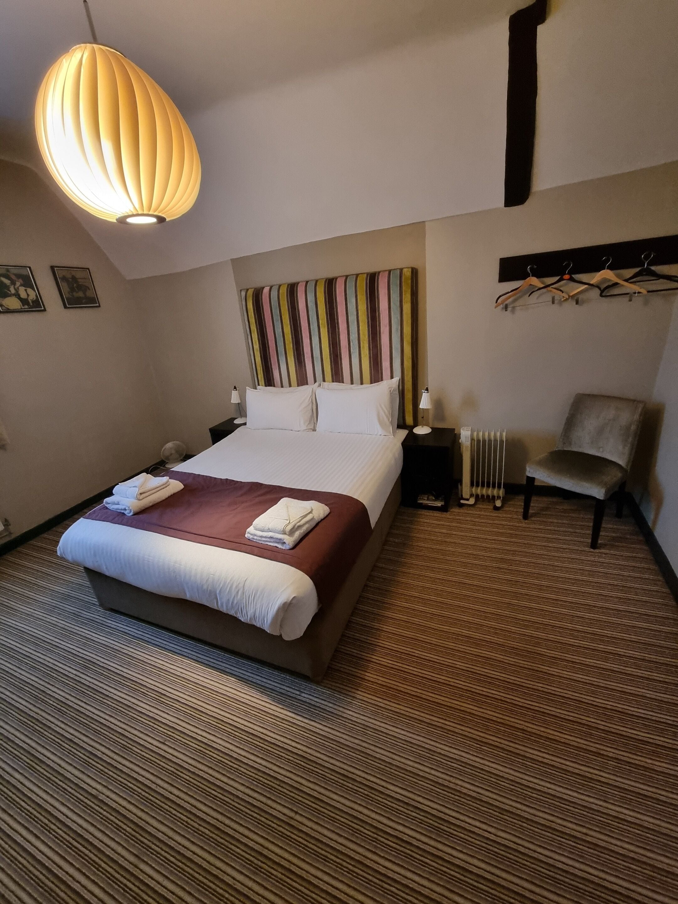 Ett hotellrum med en stor säng, en randig sänggavel, en hängande lampa, en stol och ett element.