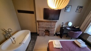 A hotel room with a large bathtub, a bed with a red and white bedspread, a flat-screen TV mounted above a brick fireplace, and a desk with a chair. - The Red Lion (Cambridge)