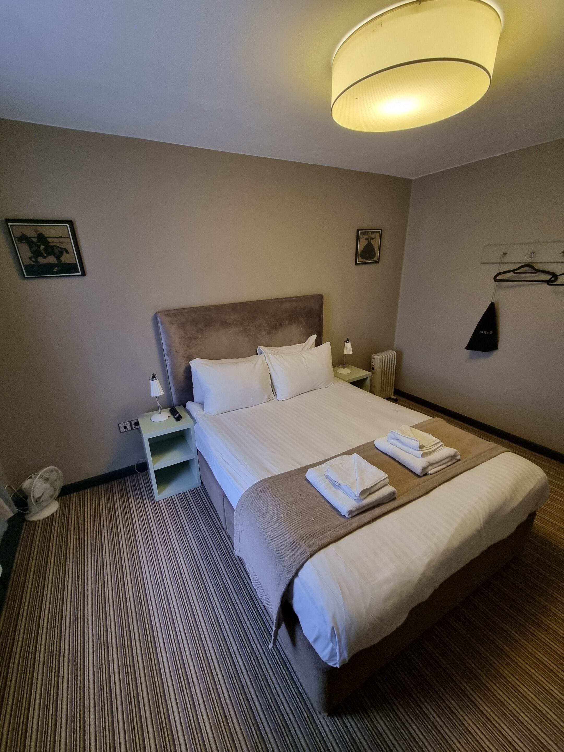 A hotel room with a large bed, a bedside table, a lamp, a fan, and a wall-mounted light fixture.