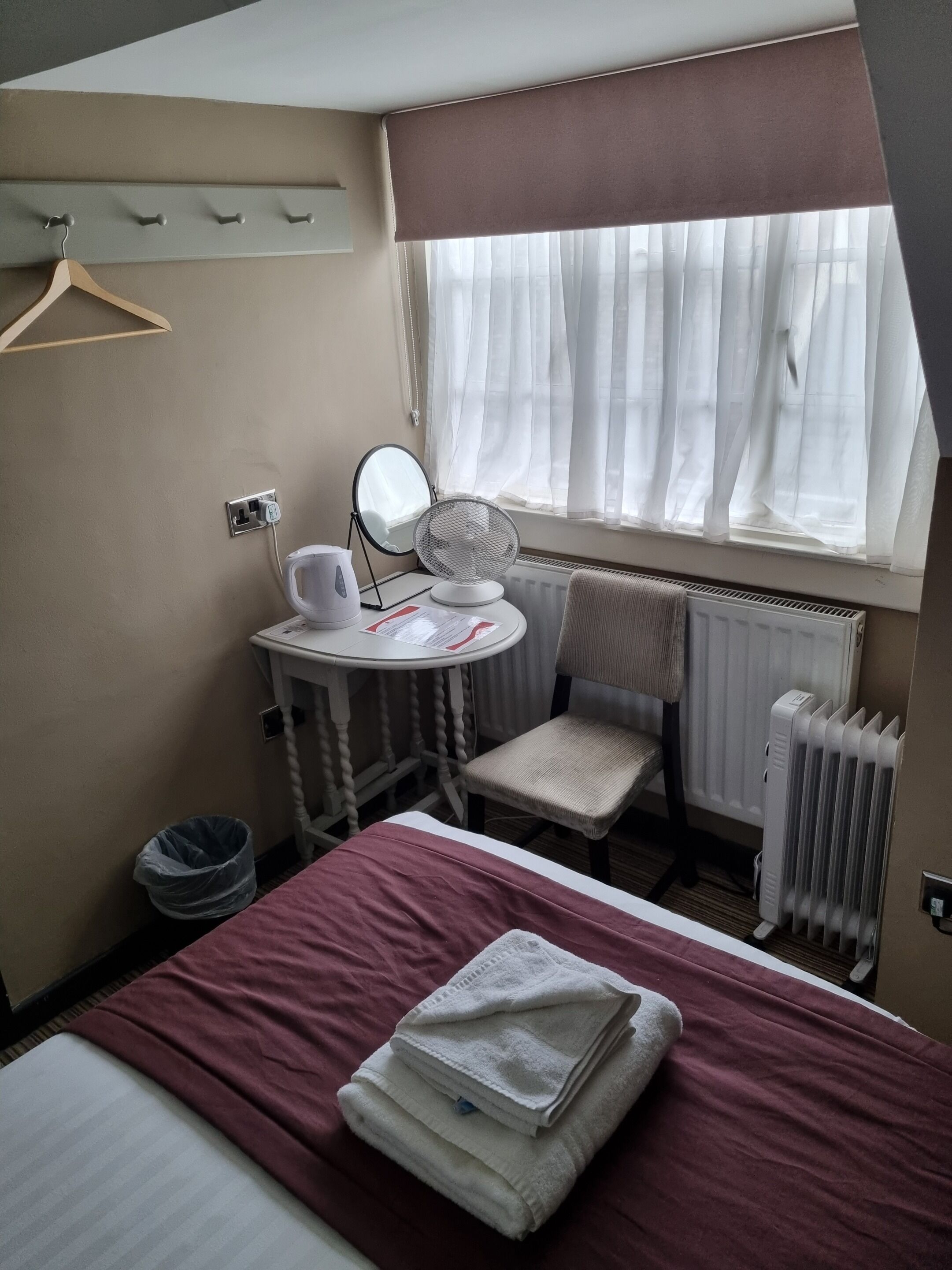Una camera d'albergo con un letto, un tavolino rotondo con uno specchio, una sedia, un termosifone e una finestra con tende leggere.