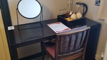 A hotel room with a desk, a chair, a trash bin, a mirror, a kettle, and a coffee maker.