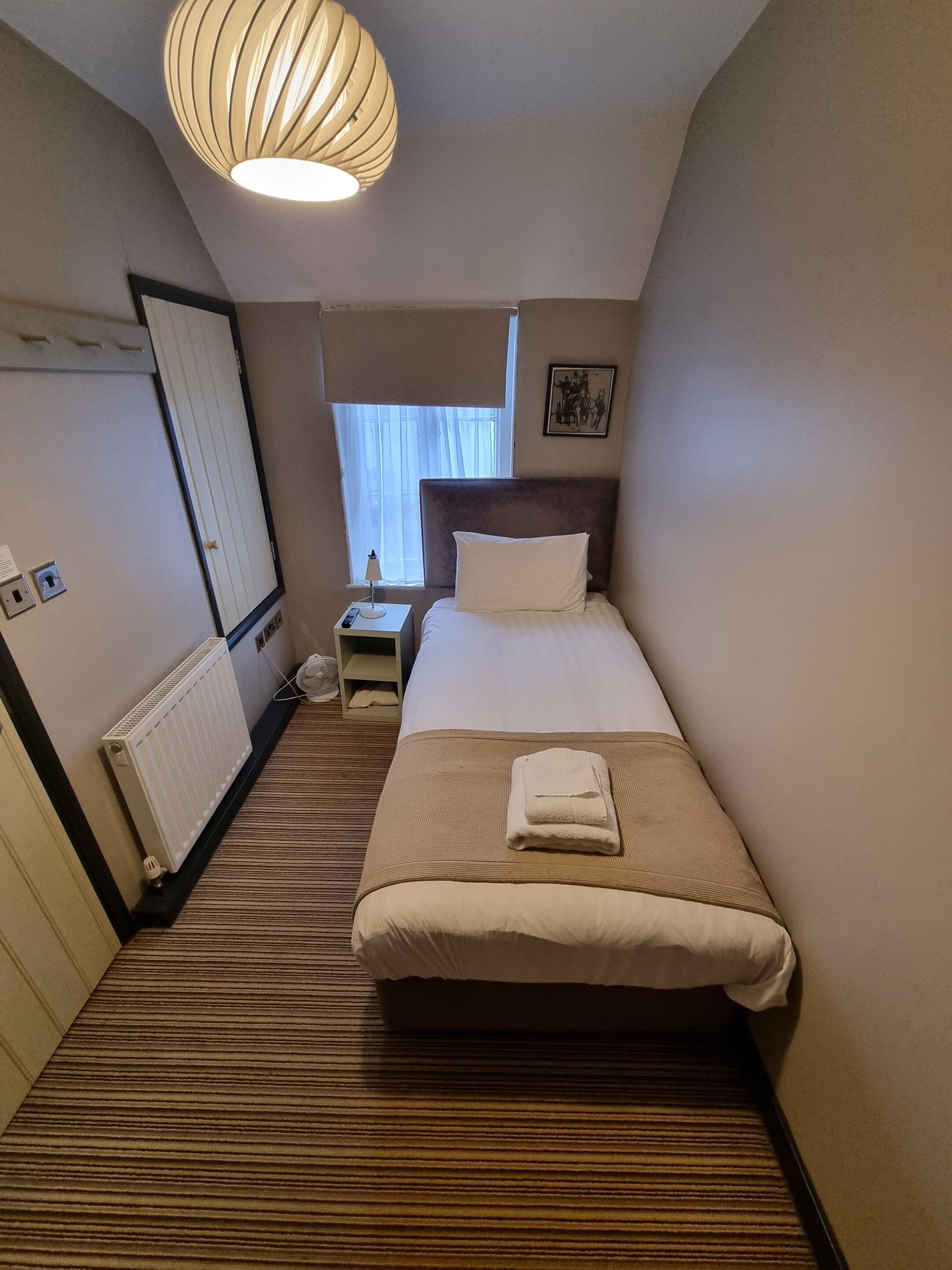 Habitación de hotel con una cama individual, una mesita de noche con lámpara, un radiador y una ventana con cortinas.