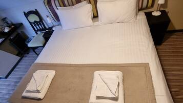 A hotel room with a neatly made bed, a bedside table with a lamp, a chair, and a small desk with a computer.