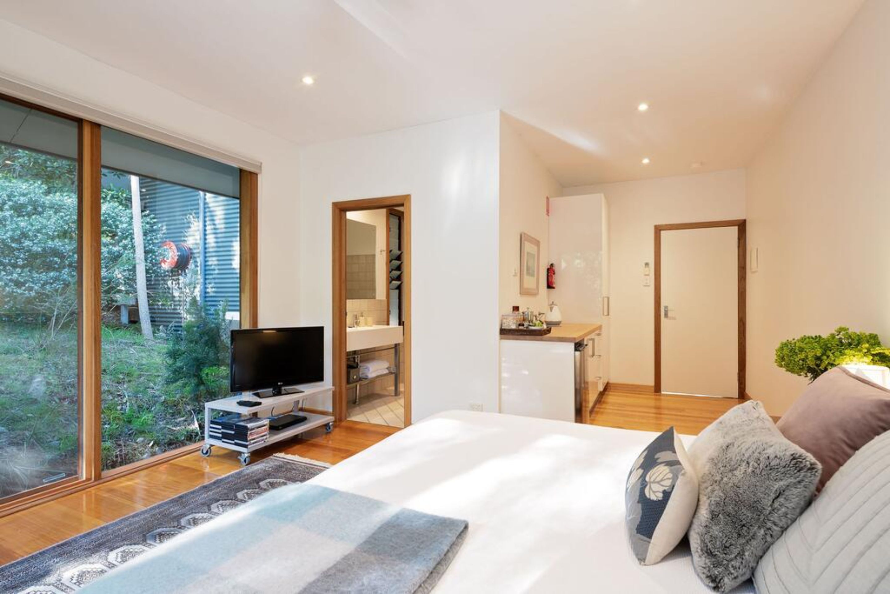 A modern bedroom with a large bed, a TV on a stand, a small kitchenette, and a sliding glass door leading to a garden.