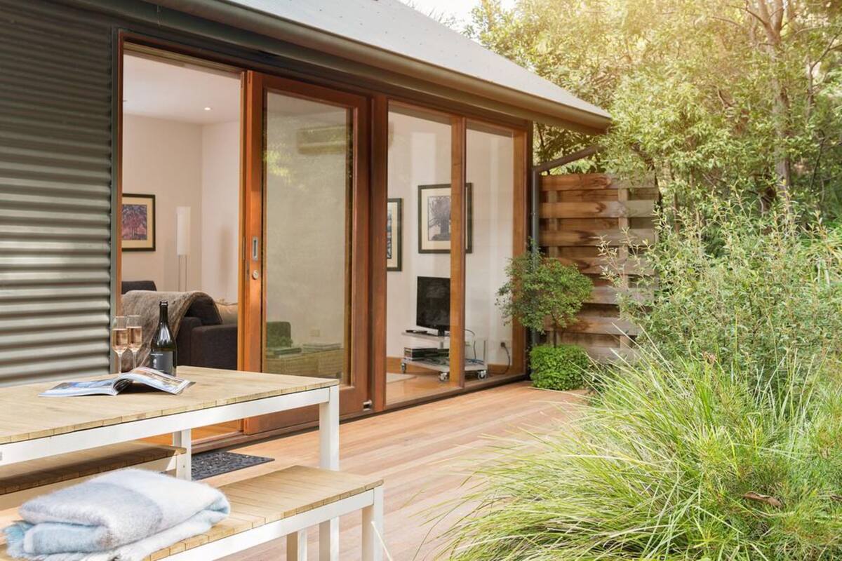A modern house with a wooden deck, sliding glass doors leading to a living area with a sofa and a TV, and a small garden area with tall grasses.