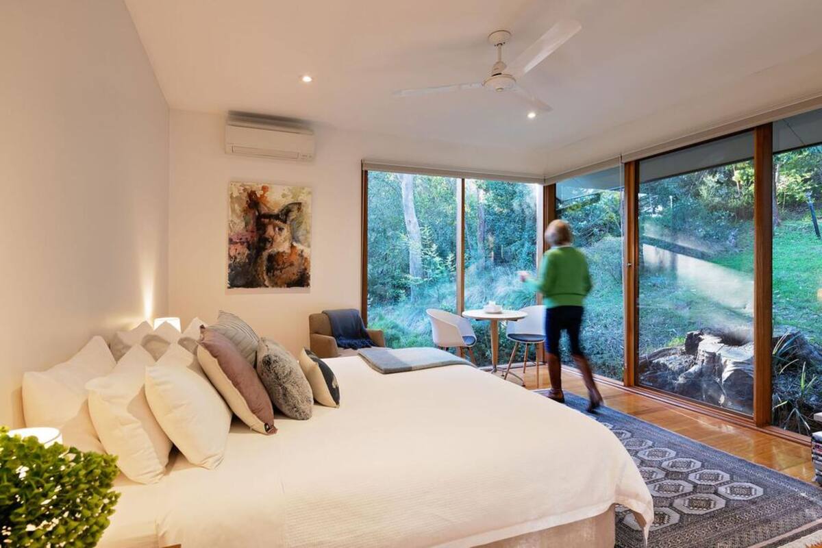 A bedroom with a large bed, a ceiling fan, and a view of the outdoors through large windows.