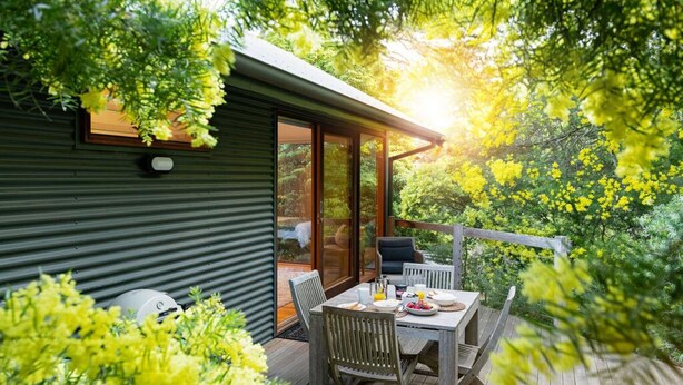 Eine Holzterrasse mit Tisch und Stühlen, umgeben von Grünpflanzen und einem Gebäude mit großen Fenstern.