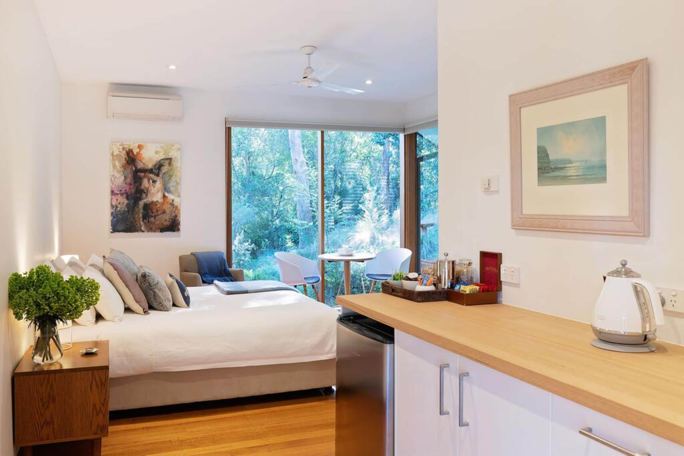 Une chambre moderne avec un lit, une table de chevet et une vue sur une forêt, visible à travers de grandes fenêtres.