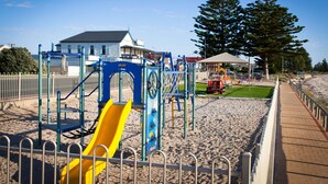 A playground with a yellow slide and climbing structures, surrounded by a sandy area and a paved walkway. - Modra's Apartments (Tumby Bay)