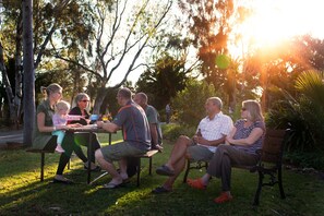A group of people seated at an outdoor table in a park with trees and sunlight in the background.