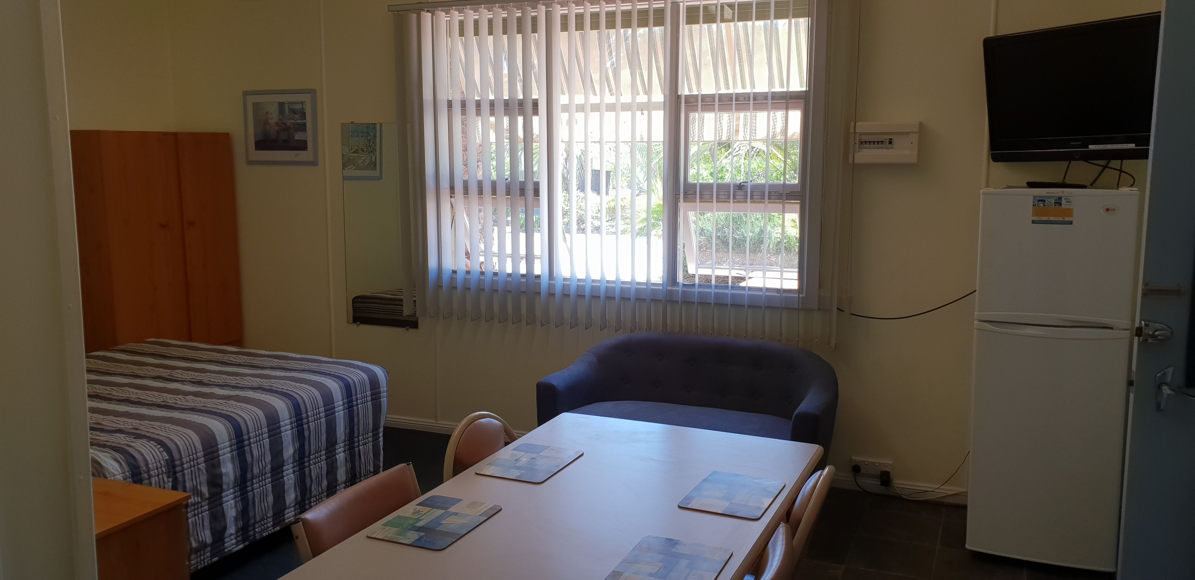 A small room with a bed, a table with chairs, a refrigerator, and a television on a stand.