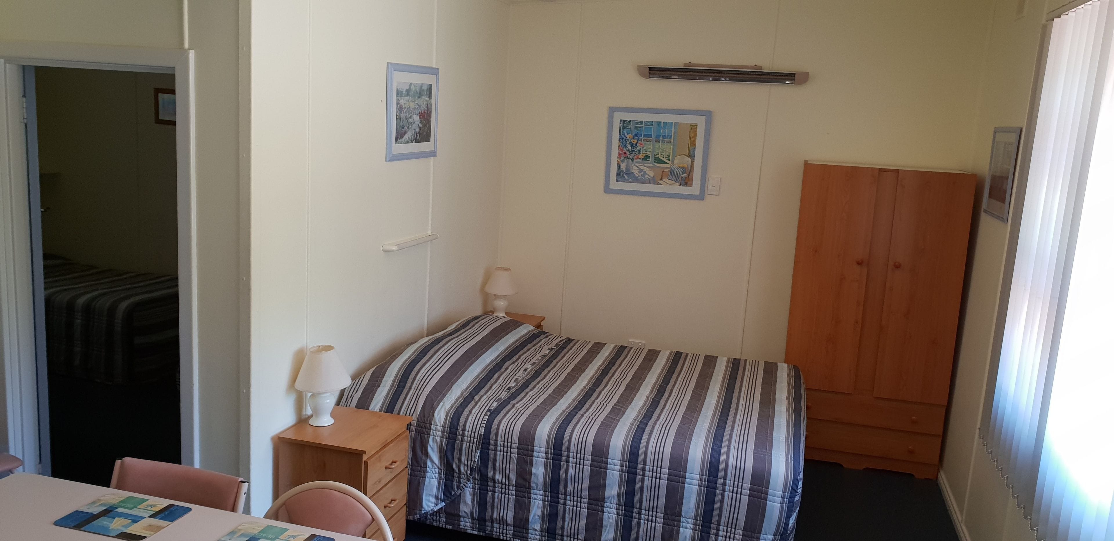 Une petite chambre à coucher, bien rangée, avec un lit simple, une armoire en bois, une table de chevet avec une lampe et deux tableaux encadrés accrochés au mur.