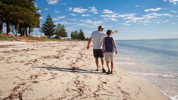 A couple walking on a sandy beach with trees and a clear sky. - Modra's Apartments (Tumby Bay)