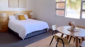 A bedroom with a bed, bedside tables, a round dining table with chairs, and a window with a view of palm trees. - Modra's Apartments (Tumby Bay)
