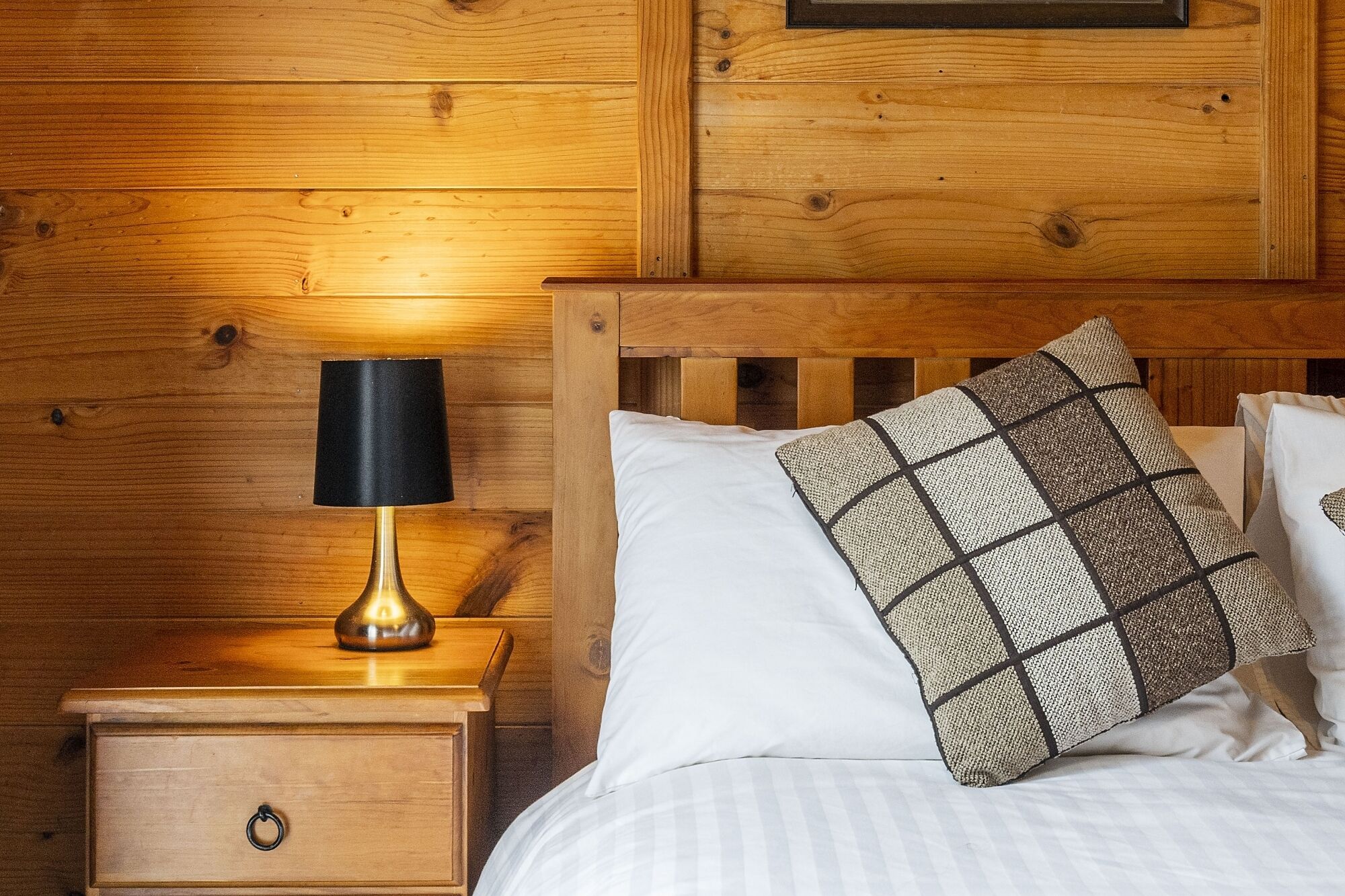 Una cama de madera con ropa de cama blanca y un cojín estampado, una lámpara de noche y una mesita de noche de madera con un cajón.