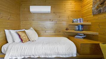 A wooden-paneled room with a bed, white bedding, a bookshelf, and an air conditioning unit.
