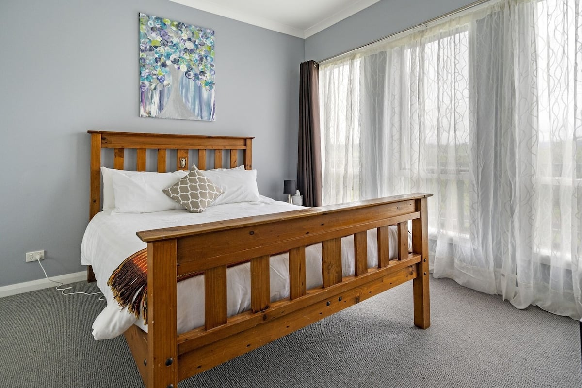 Un dormitorio con cama de madera, ropa de cama blanca, un cuadro en la pared y cortinas translúcidas.