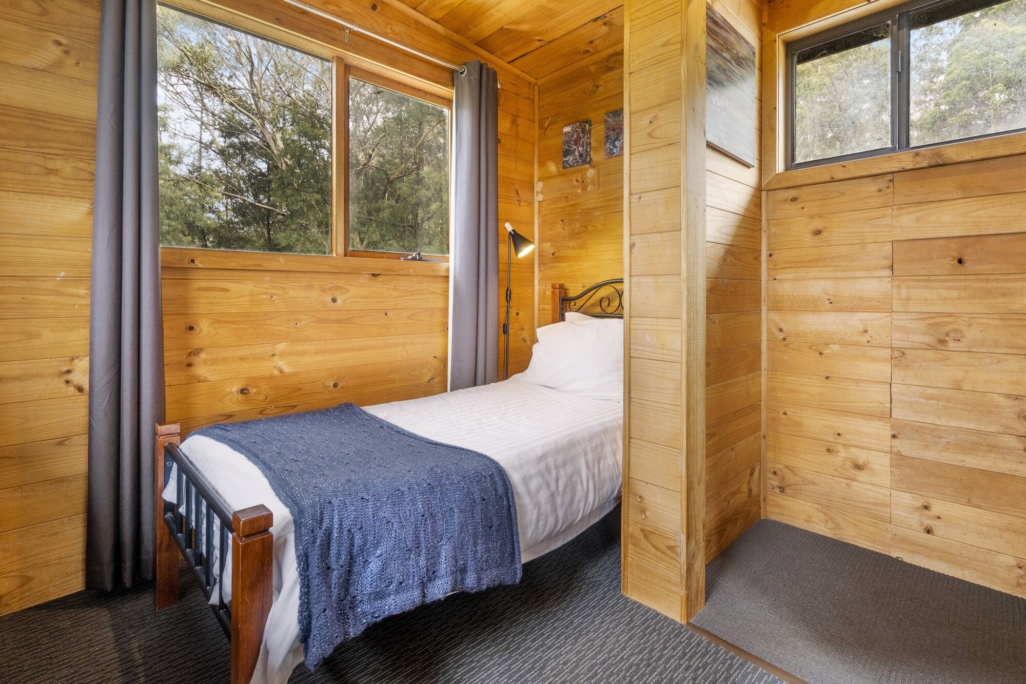 A small wooden cabin bedroom with a single bed, a nightstand, a lamp, and a window with curtains.