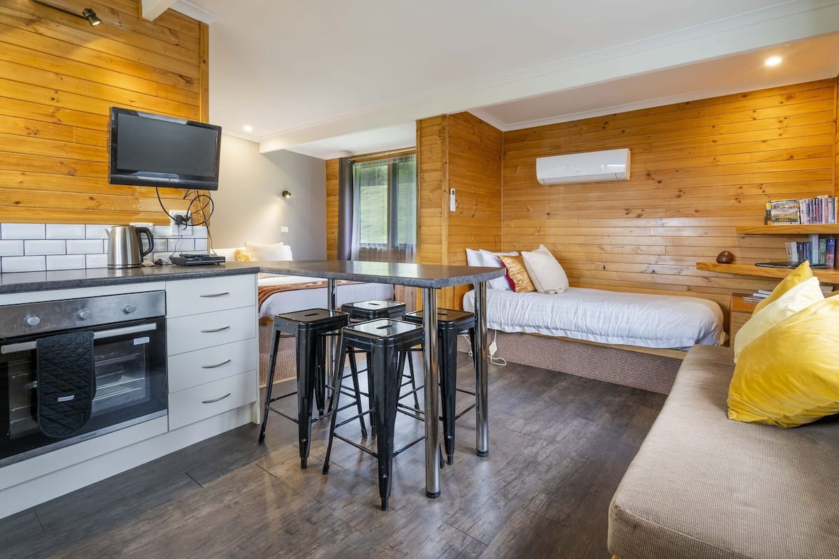 Una habitación compacta y moderna con kitchenette, una zona de comedor con taburetes, cama y estantería.