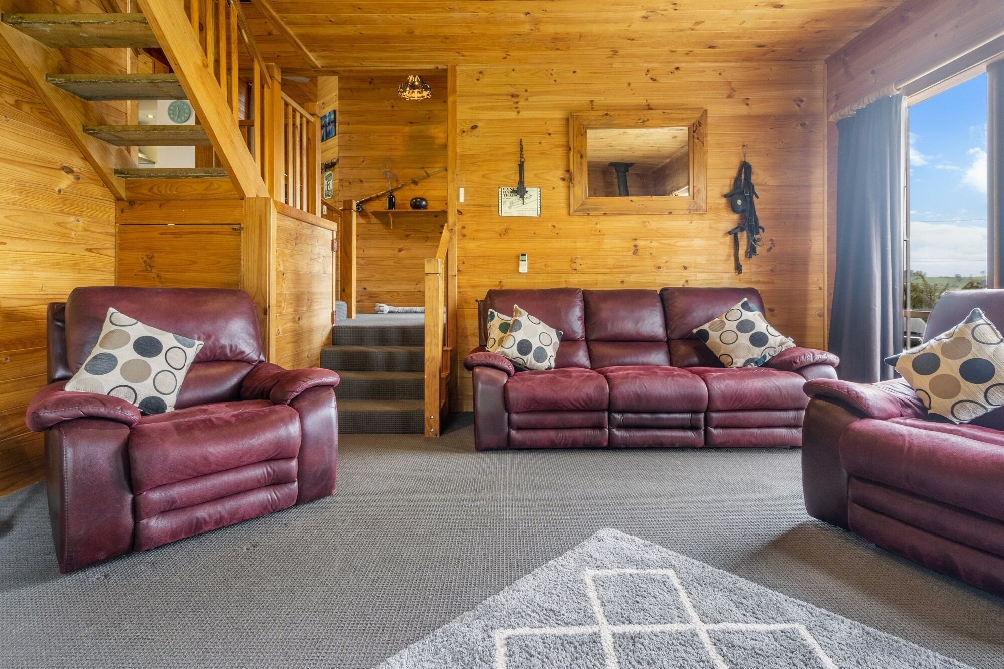 L’intérieur chaleureux d’un chalet avec un escalier en bois, des canapés en cuir ornés de coussins à motifs et une grande fenêtre donnant sur le paysage extérieur.