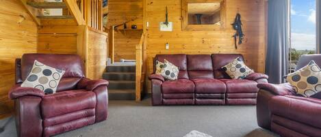 L’intérieur chaleureux d’un chalet avec un escalier en bois, des canapés en cuir ornés de coussins à motifs et une grande fenêtre donnant sur le paysage extérieur.