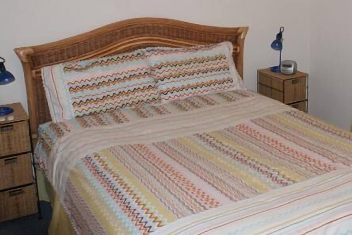 A bed with a patterned bedspread and pillows, a wooden headboard, and a bedside table with a lamp.