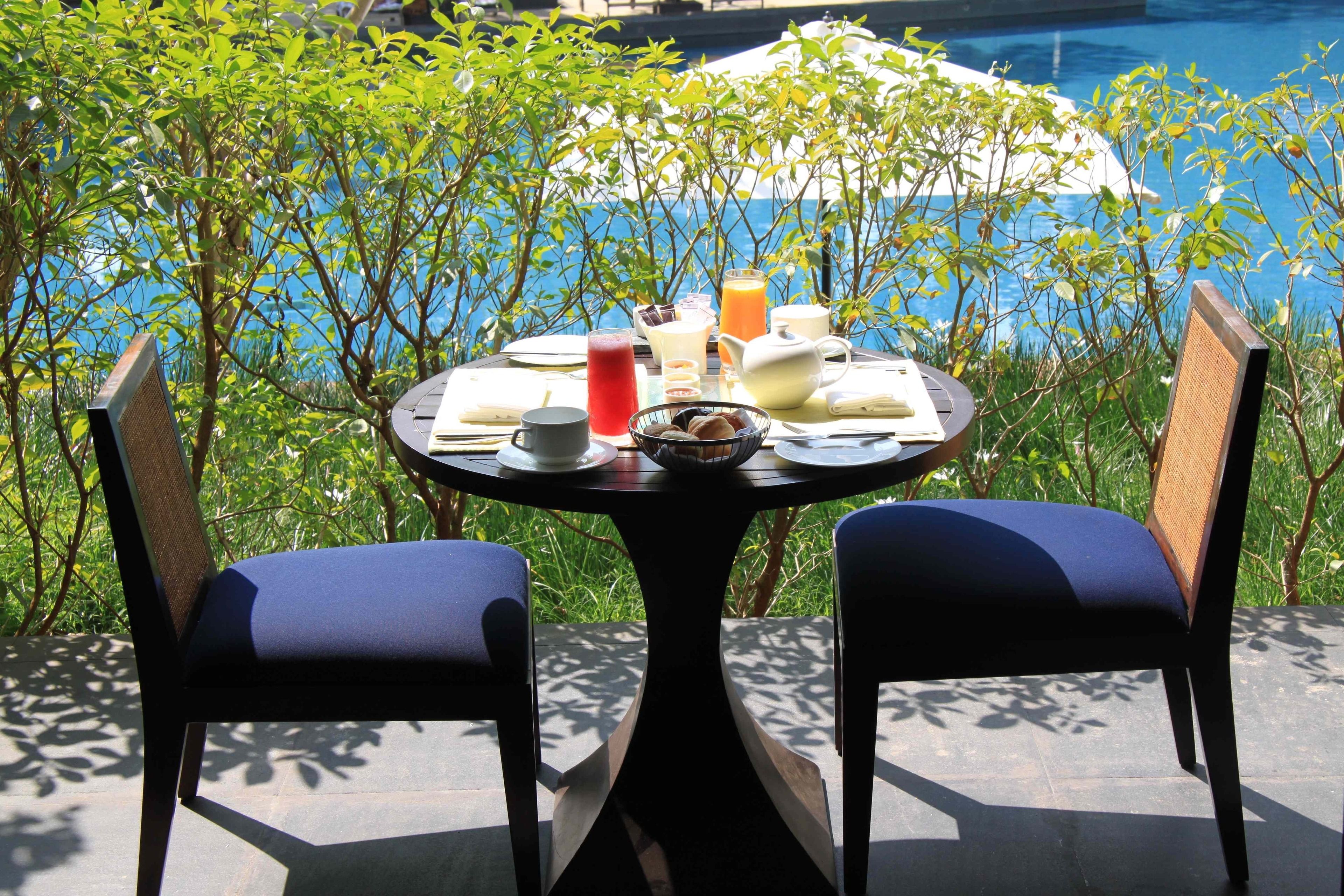 A round table with two chairs, a bowl of cookies, a pitcher, and a glass of orange juice, set against a poolside view.