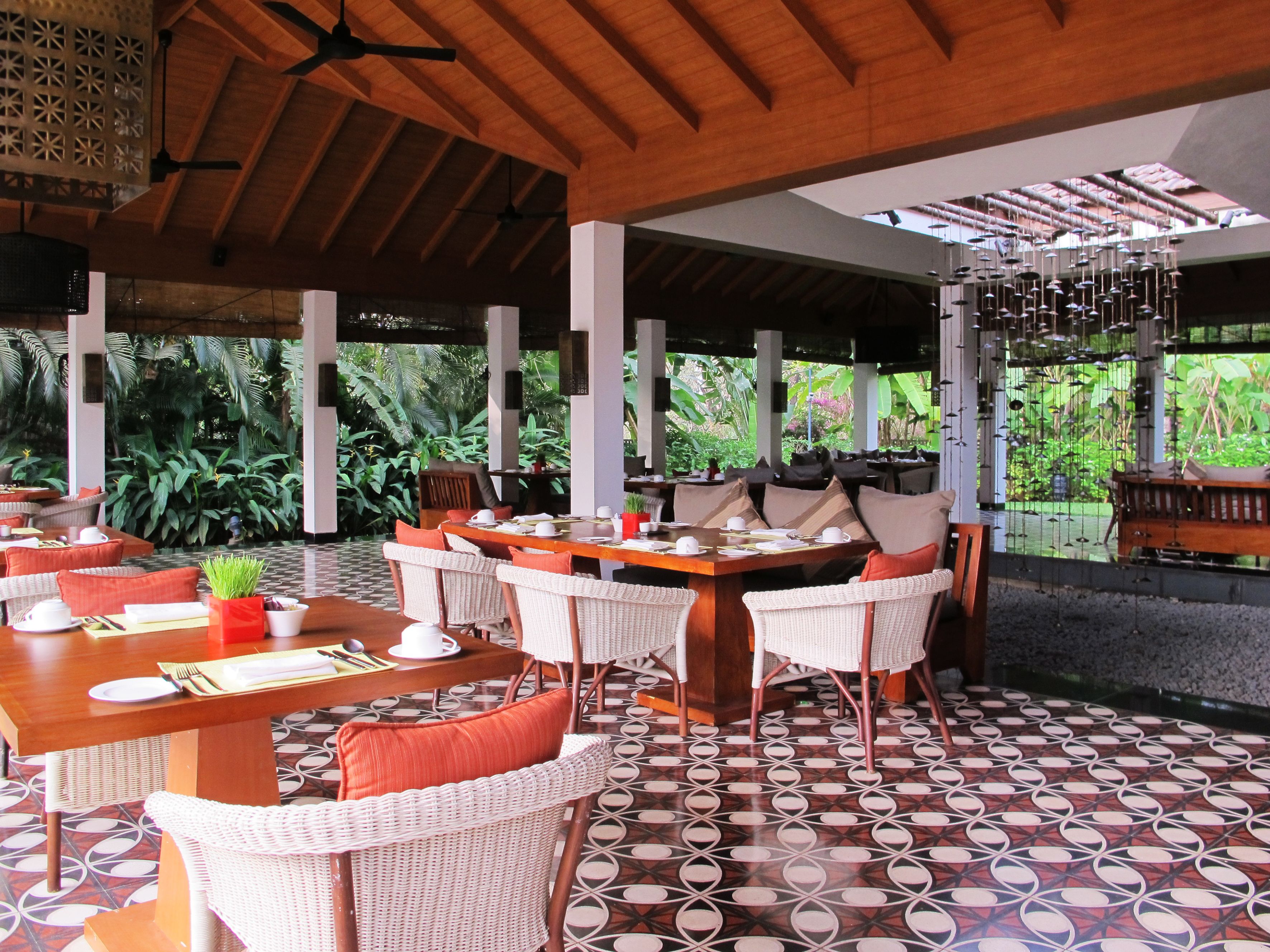 A restaurant with outdoor seating, covered by a wooden roof, surrounded by lush greenery.