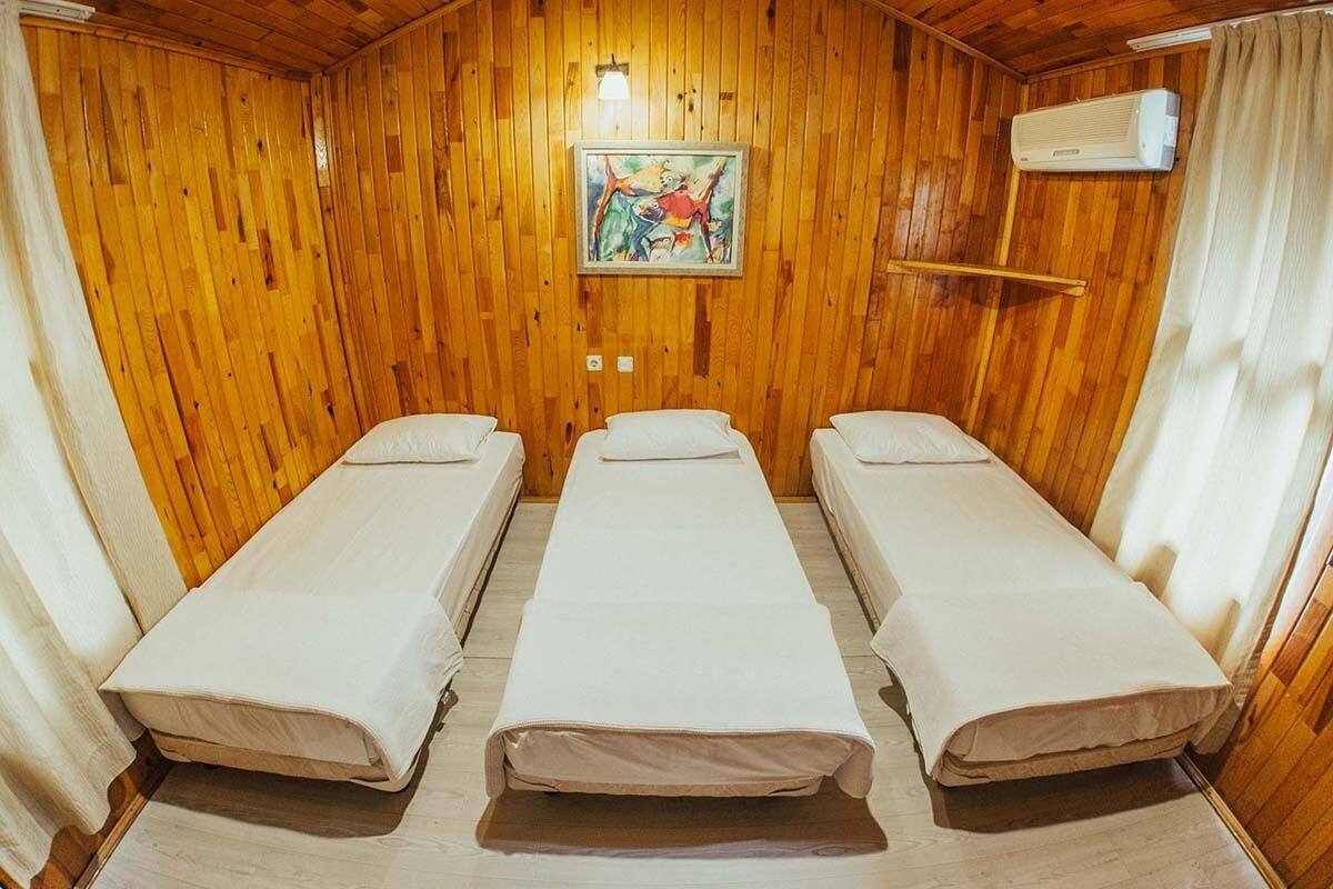 A room with three single beds aligned in a row, wooden walls, a ceiling light, and an air conditioning unit.