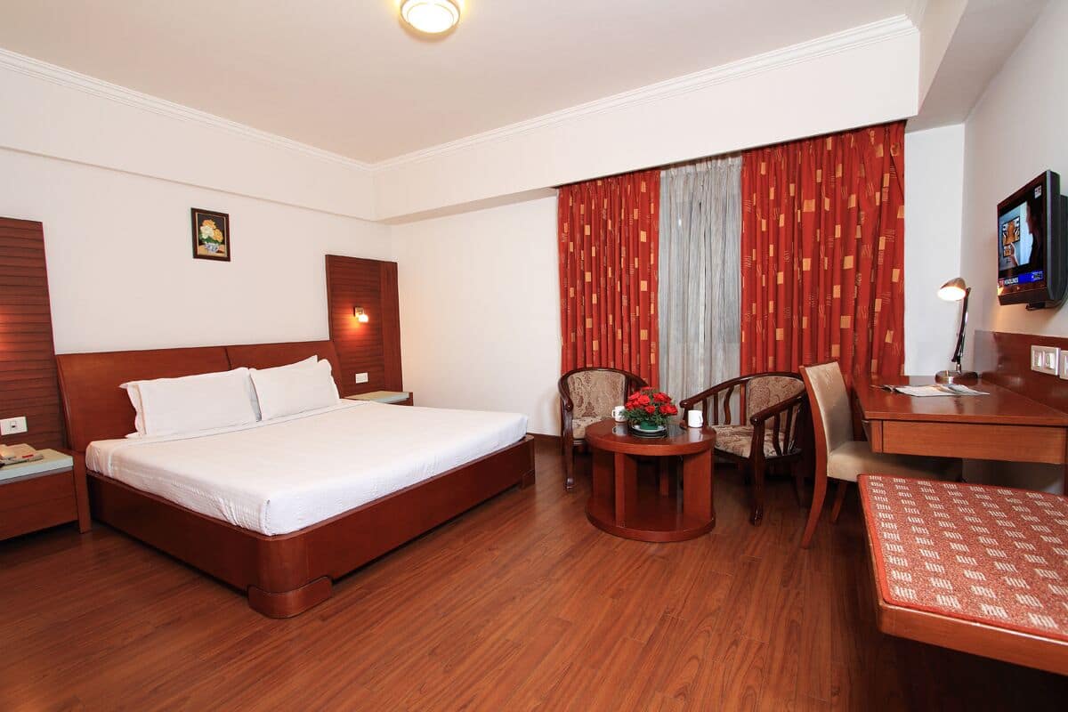 Quarto de hotel com uma cama grande, uma mesa de escritório com cadeira, uma mesinha com flores e uma televisão na parede.