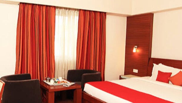A hotel room with a large bed, two armchairs, a small table, and a window with red curtains. -  Parijatha Gateway (Bengaluru)