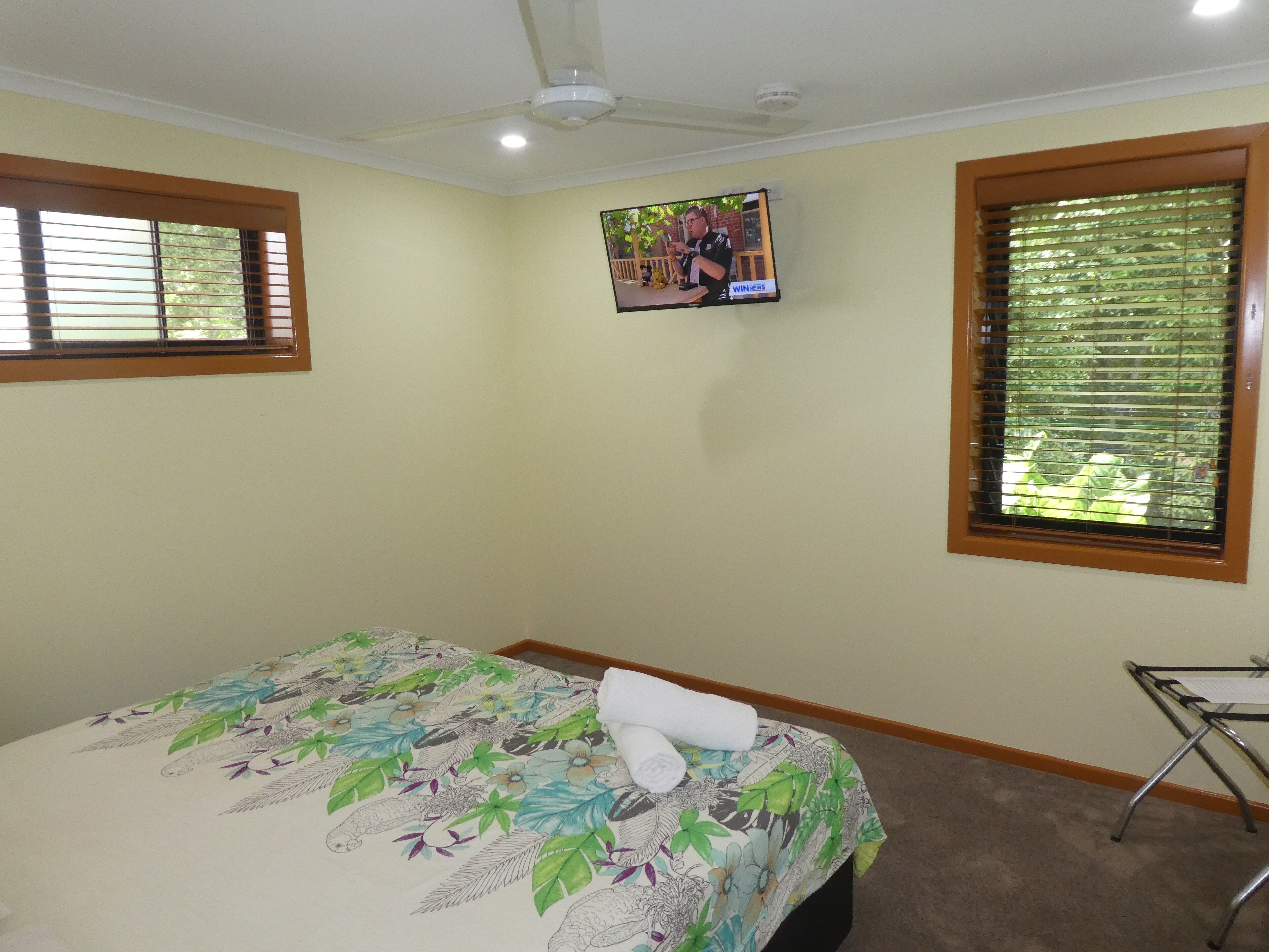 A bedroom with a bed, a TV mounted on the wall, and a window with blinds.