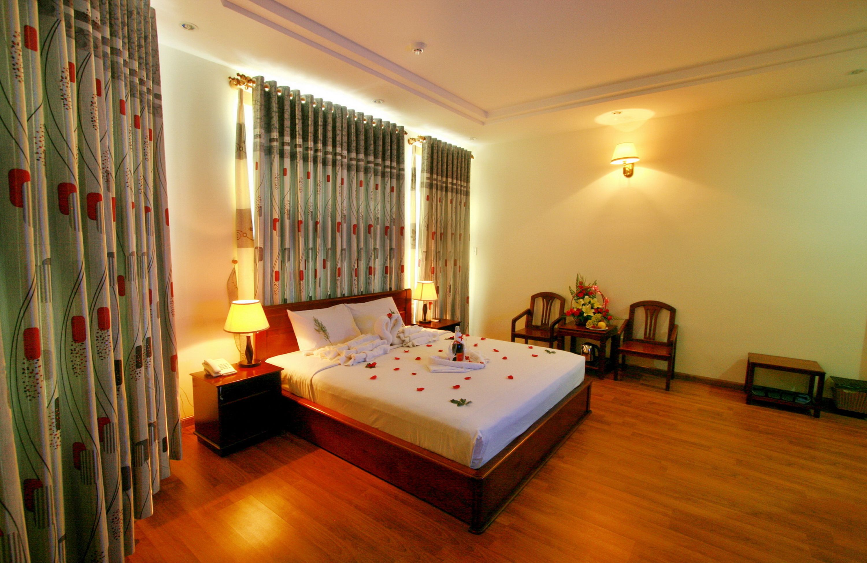 A neatly made bed with a white bedspread and pillows, a bedside table with a lamp, and a chair with a vase of flowers.