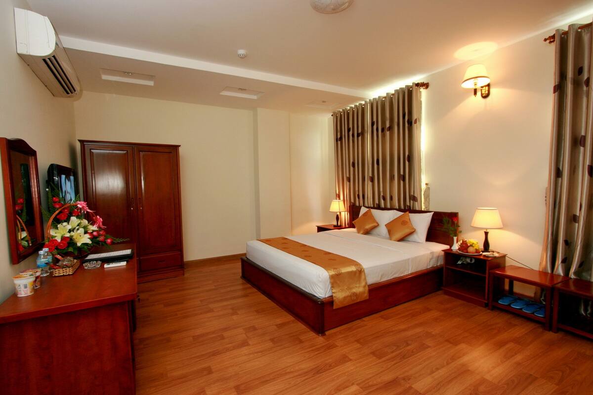 Ein Hotelzimmer mit Holzboden, einem großen Bett mit weißer und brauner Bettdecke, zwei Nachttischen mit Lampen, einem Holzschrank und einem Spiegel an der linken Wand.
