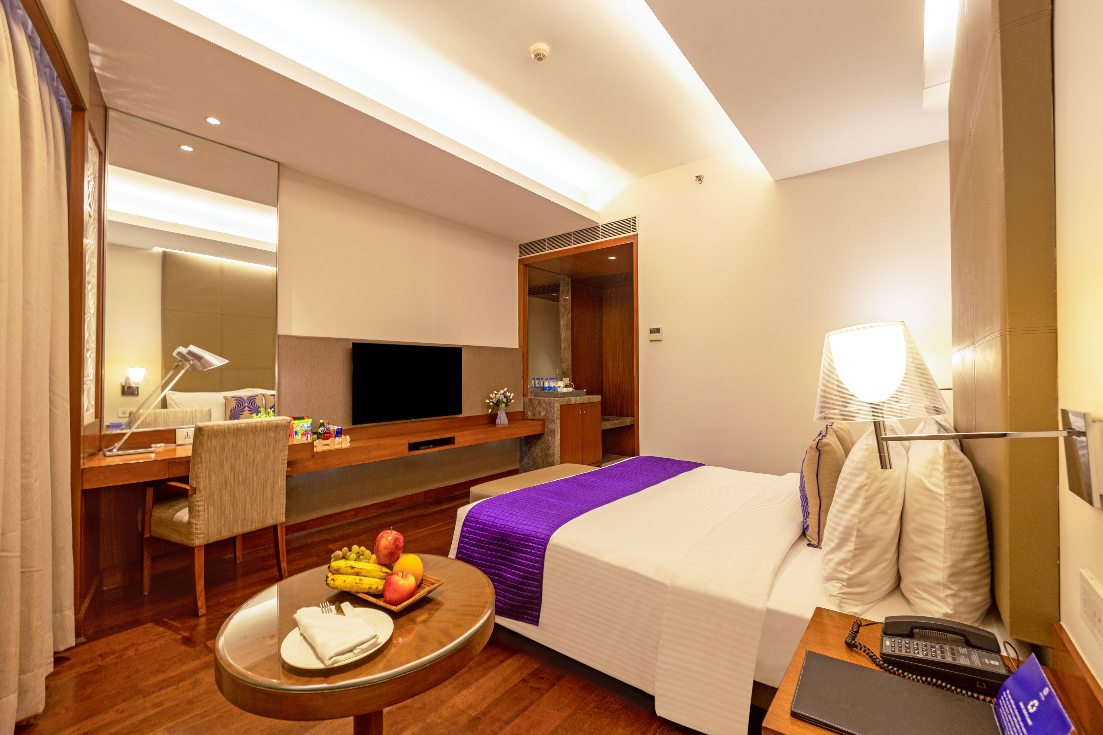 A hotel room with a bed, bedside table, desk, chair, and a small round table with fruits and a plate.