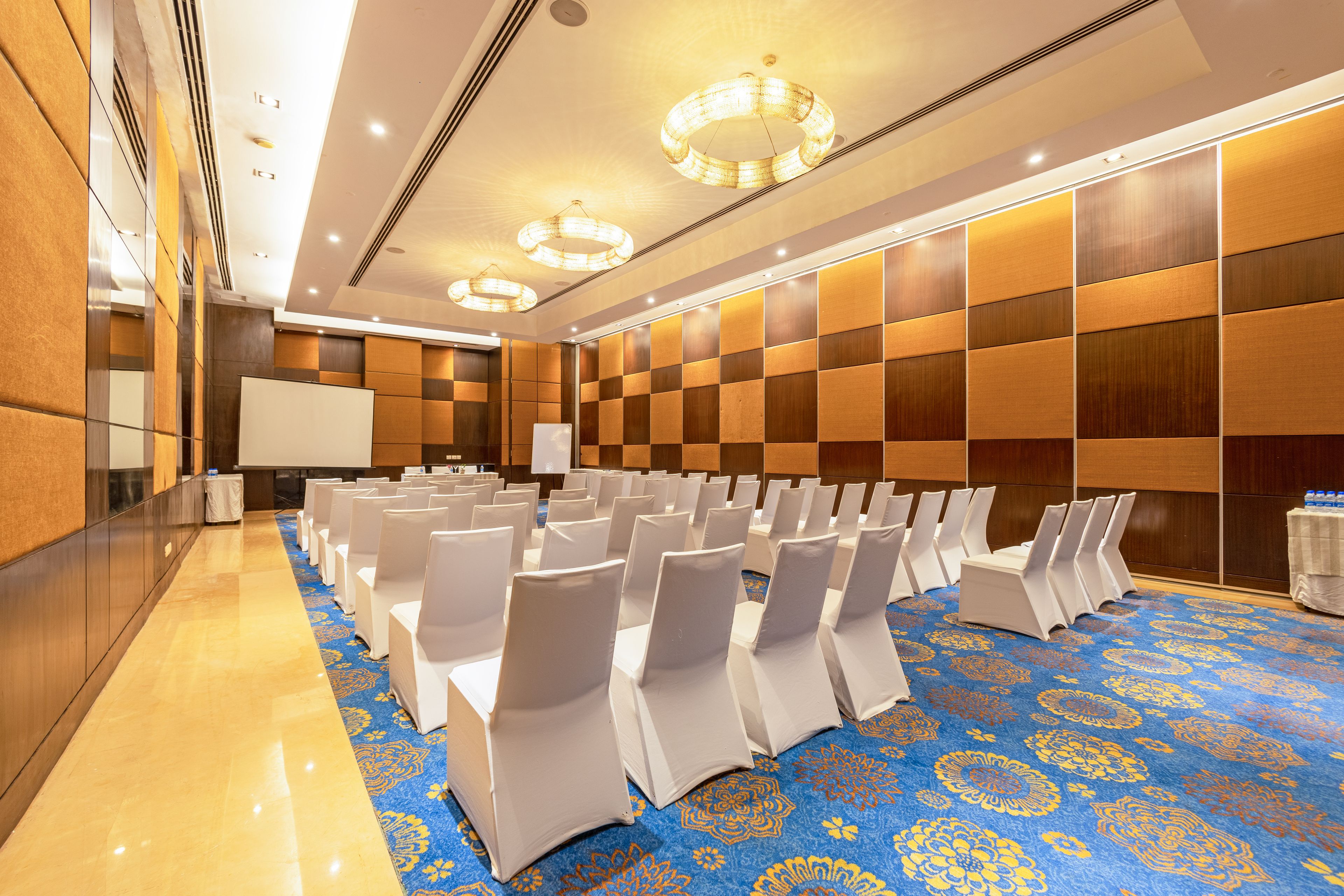 A conference room with rows of white chairs, a large screen, and a blue patterned carpet.