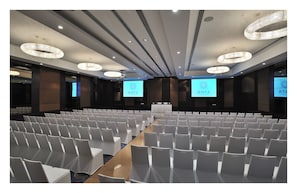 Une salle de conférence avec des rangées de chaises blanches, une longue table et de grands écrans affichant le logo « ANYA ».