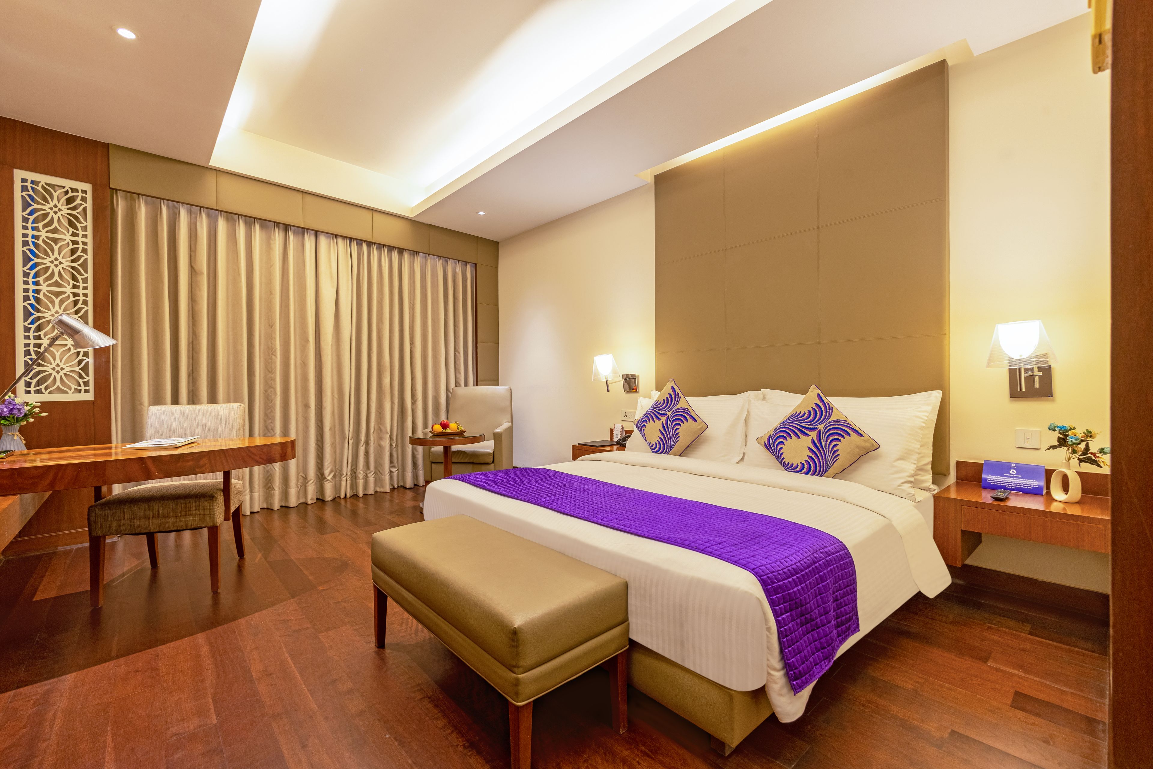 A hotel room with a bed, a desk, a chair, a small table with fruit, and a bench.