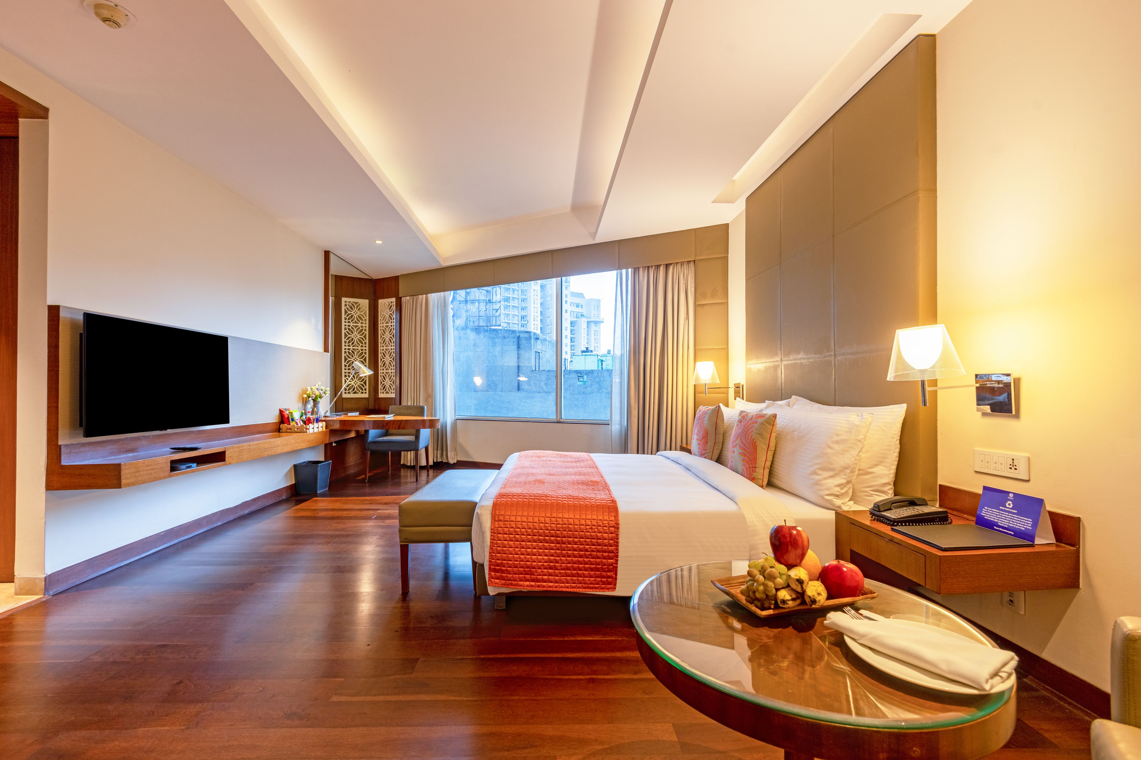 A hotel room with a bed, a desk, a TV, a glass table with fruit, and a view of the city.