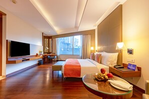 Une chambre d’hôtel avec un lit, un bureau, une télévision, une table en verre avec des fruits, et une vue sur la ville.