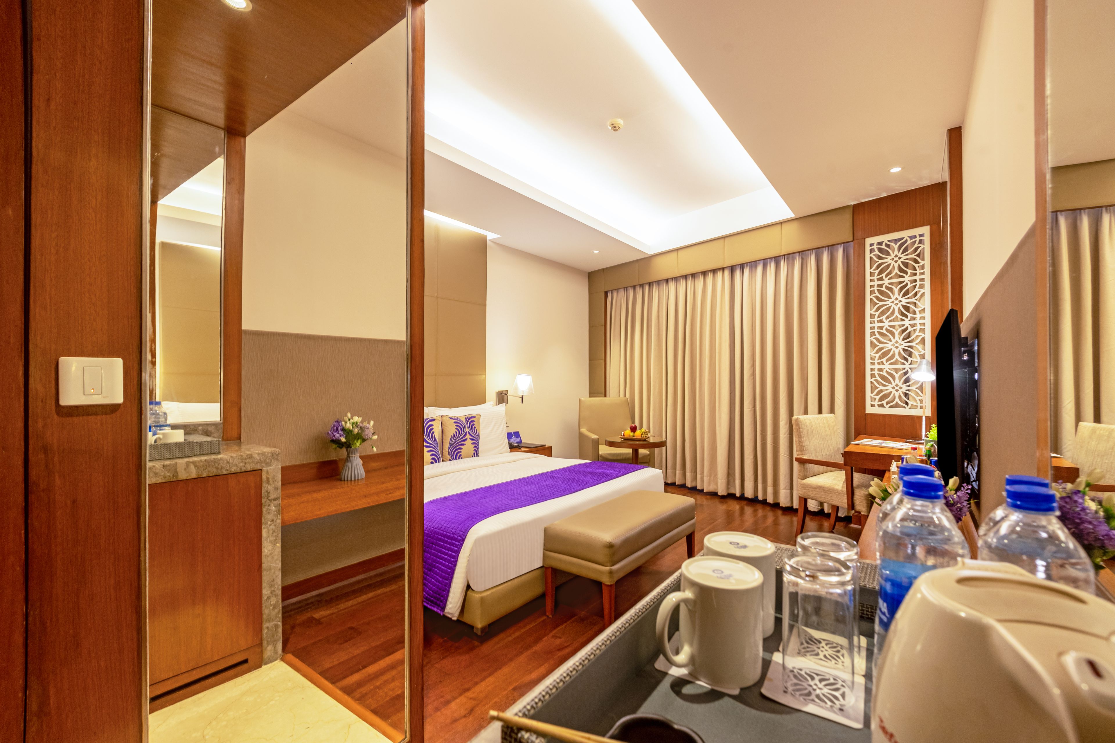 A hotel room with a bed, a desk, a chair, a TV, and a bathroom visible through a glass partition.