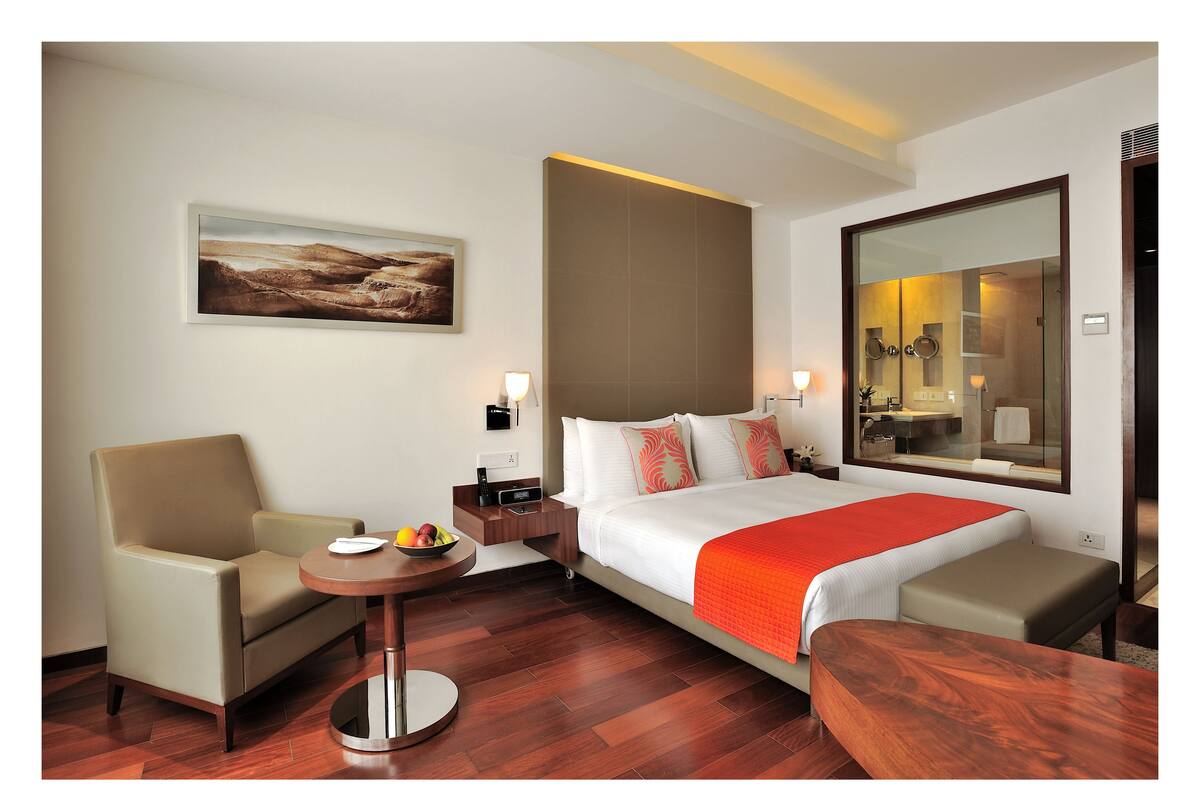 A hotel room with a bed, a chair, a small table with fruit, a painting on the wall, and a large mirror.